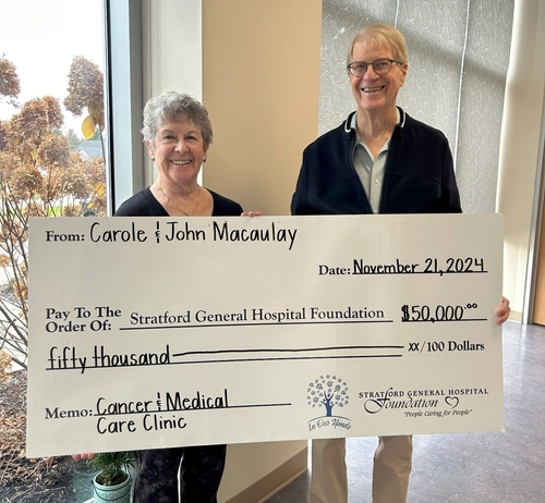 Carole and John Macaulay smile while holding a large ceremonial cheque for $50,000 made out to the Stratford General Hospital Foundation. The cheque is dated November 21, 2024, and designated for the Cancer & Medical Care Clinic. The photo is taken inside the Foundation office, with a window showing trees in the background.